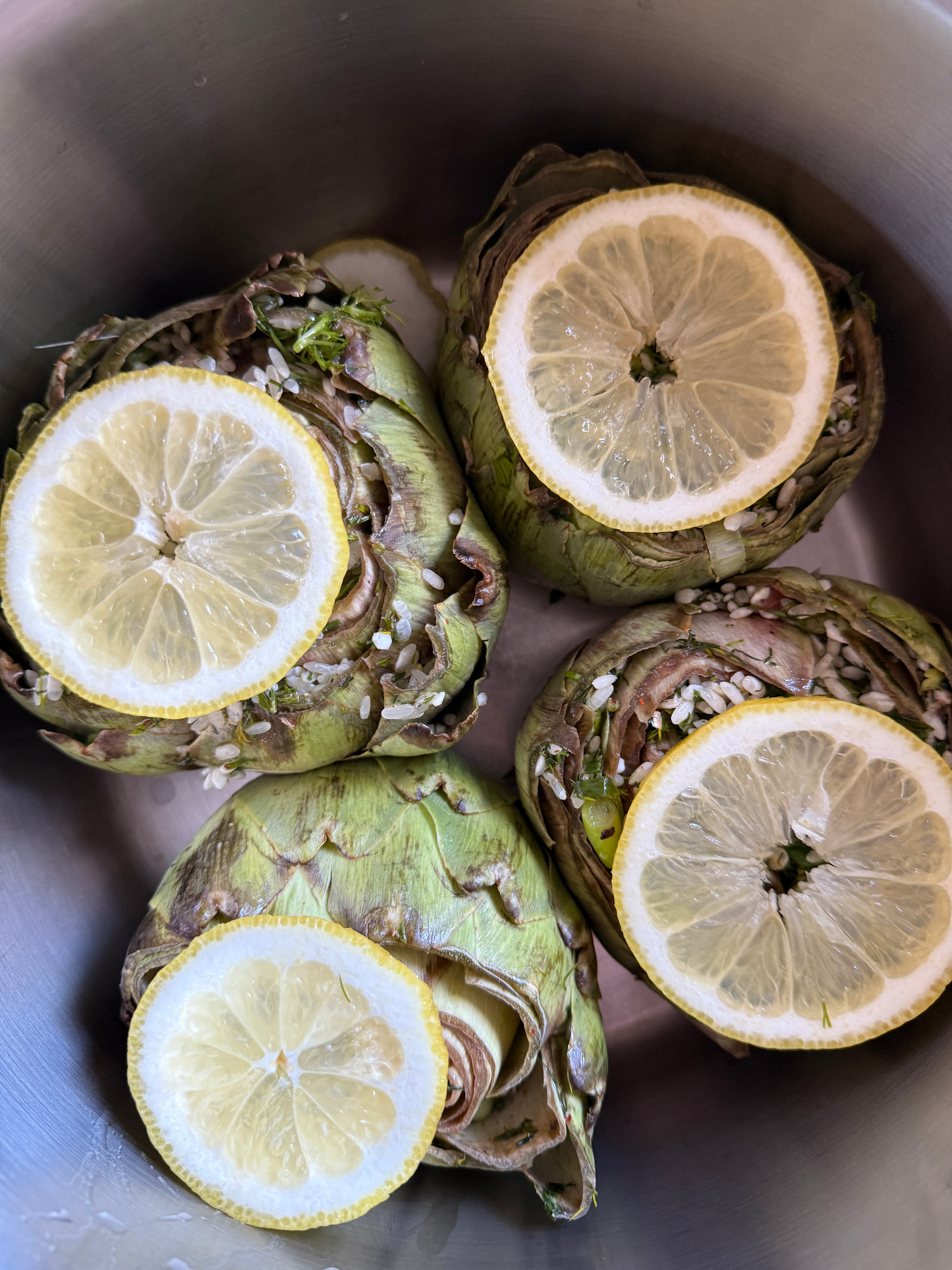 Vier geputzte Artischocken mit grün-violetten Blättern, belegt mit Zitronenscheiben und gehacktem Knoblauch, in einem Edelstahltopf.