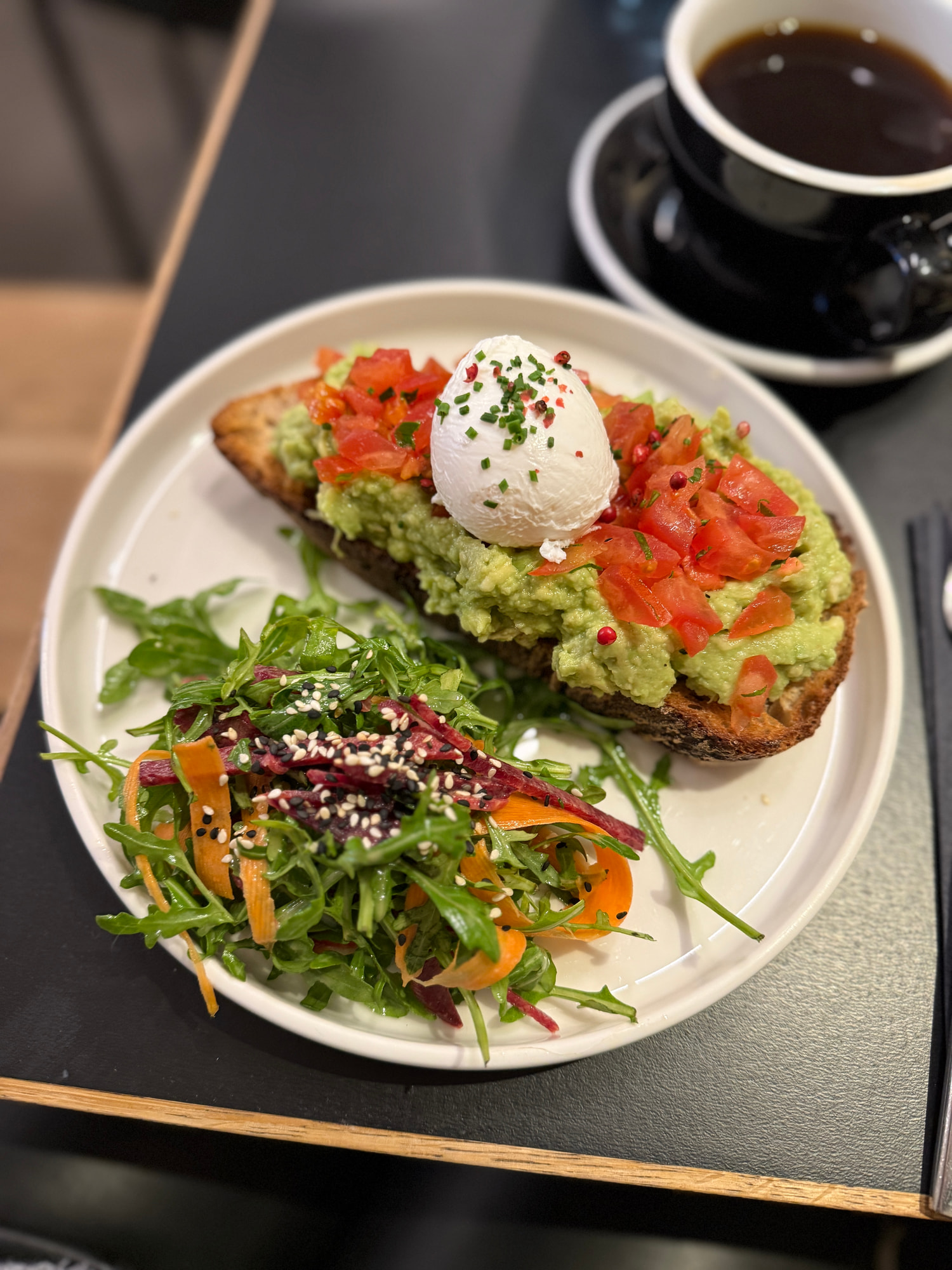 Avocadobrot mit pochiertem Ei, Tomatenwürfeln und Schnittlauch, daneben ein Rucolasalat mit Paprikastreifen, dahinter eine Tasse schwarzer Kaffee.