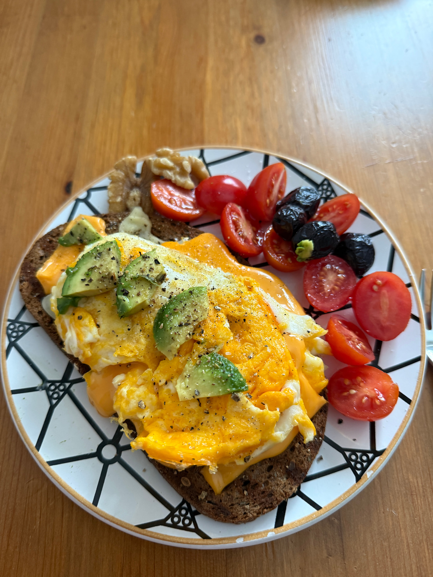 Rührei mit Avocadoscheiben auf Vollkornbrot, dazu halbierte Cocktailtomaten, schwarze Oliven und Walnüsse auf einem schwarz-weiß gemusterten Teller.