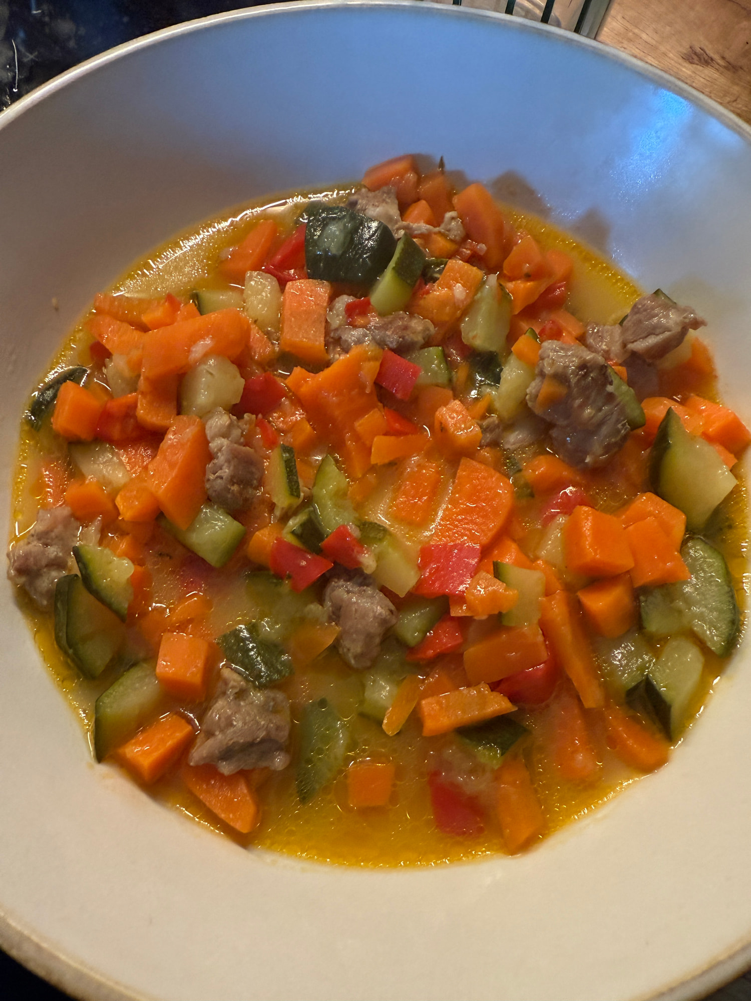 Ein tiefer heller Teller mit Gemüseeintopf aus Karotten, Zucchini, roter Paprika und kleinen Fleischstücken in goldgelber Brühe.