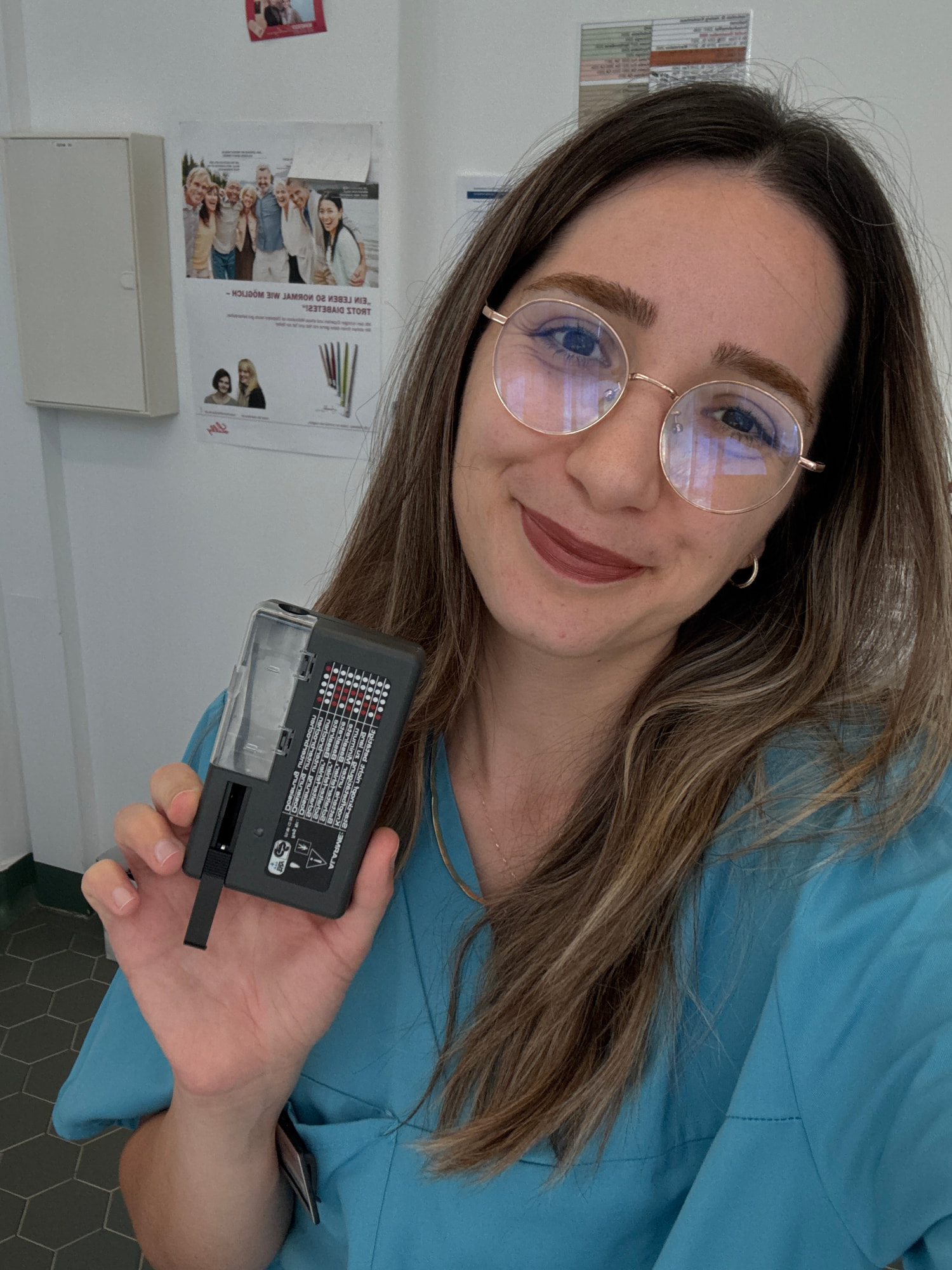 Elif Çimen Güngör hält in türkisfarbener Berufskleidung und mit Brille ein kleines schwarzes Gerät mit Display in die Kamera, im Hintergrund hängen Plakate.