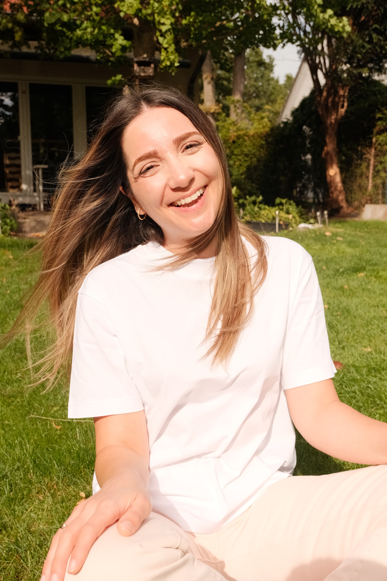 Elif Çimen Güngör sitzt lachend im weißen T-Shirt auf einer Wiese im Garten, hinter ihr Bäume und ein Haus in der Sonne.