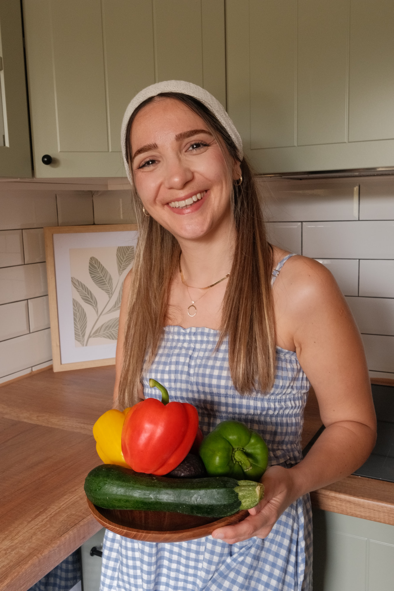 Elif Çimen Güngör hält in einer Küche eine Holzschale mit roter, gelber und grüner Paprika, Zucchini und Avocado und lächelt in die Kamera.