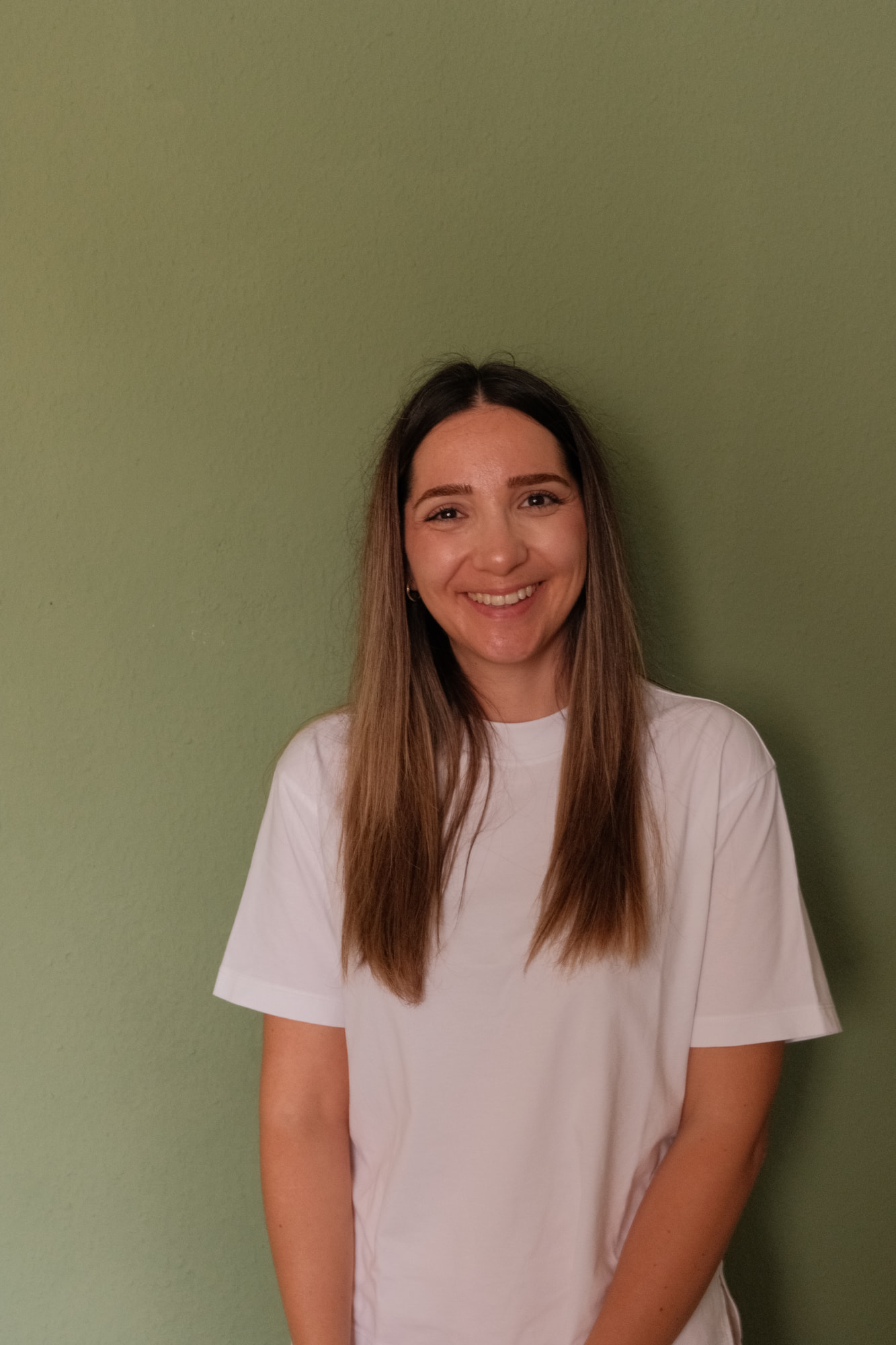 Elif Çimen Güngör steht lächelnd in einem weißen T-Shirt vor einer olivgrünen Wand, die langen Haare fallen über die Schultern.