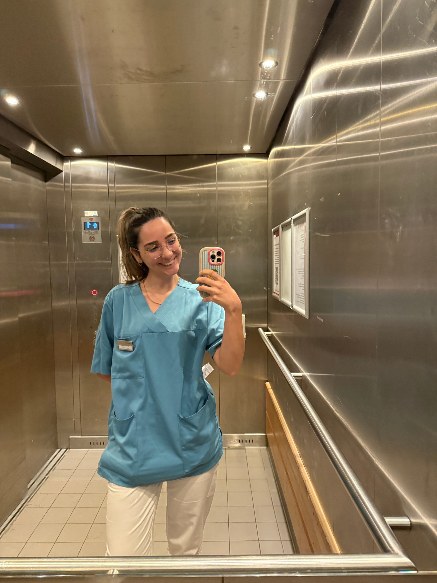Elif Çimen Güngör in türkisfarbener Berufskleidung und weißer Hose fotografiert sich lächelnd im Spiegel eines Aufzugs aus Edelstahl.