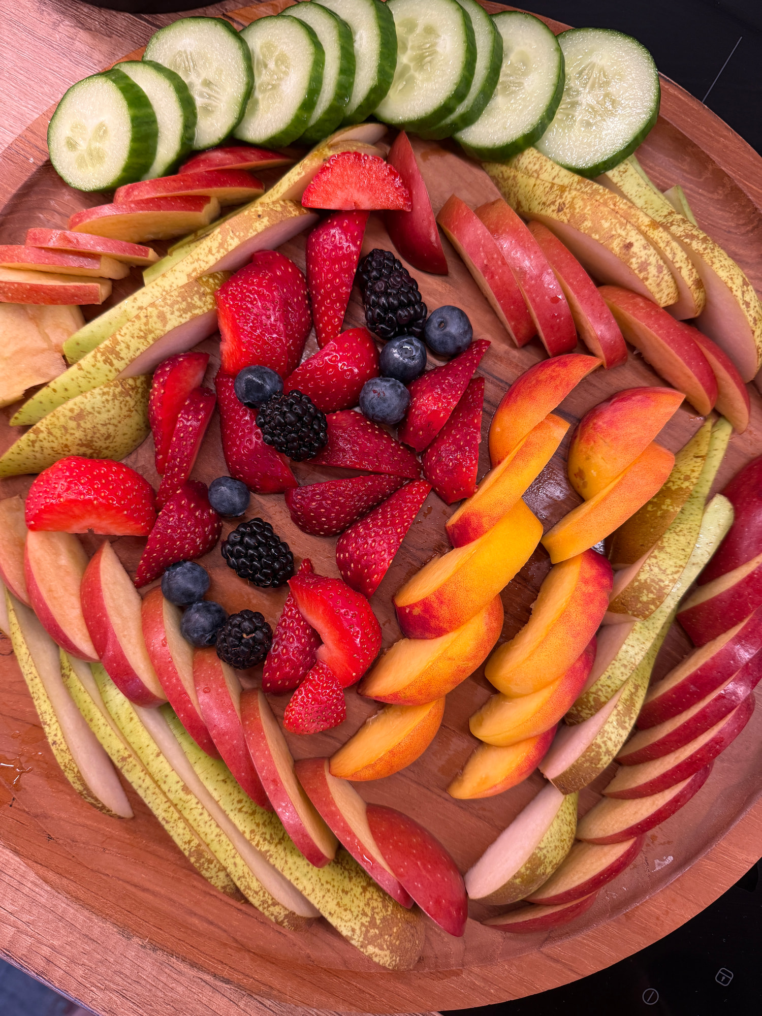 Eine Holzplatte mit Gurkenscheiben, roten und grünen Apfelspalten, Erdbeeren, Brombeeren, Heidelbeeren und Nektarinenspalten, kreisförmig angerichtet.