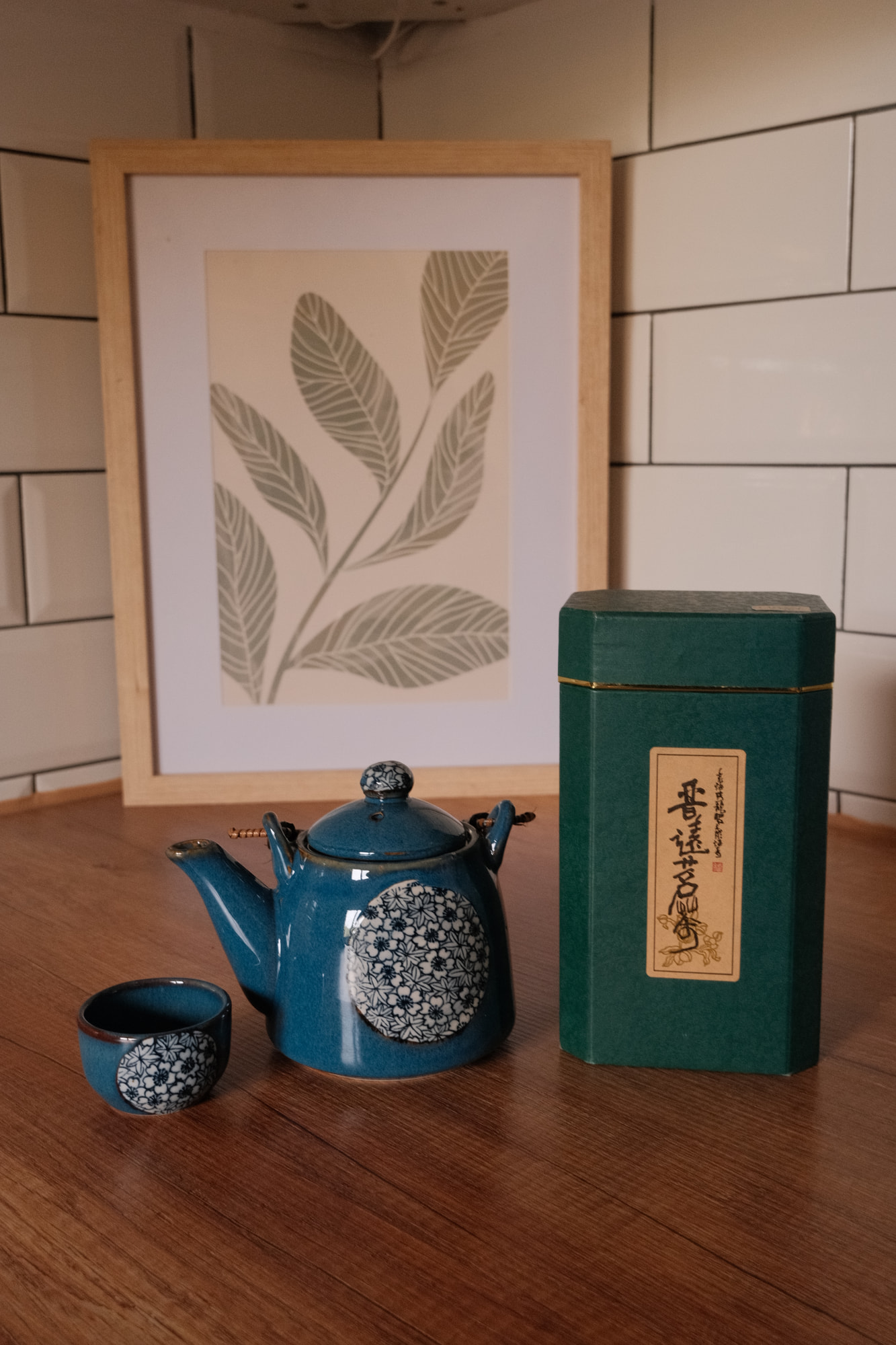 Eine petrolfarbene Teekanne mit weißem Blütenmuster und eine passende kleine Tasse stehen neben einer dunkelgrünen Teedose auf einem Holztisch.