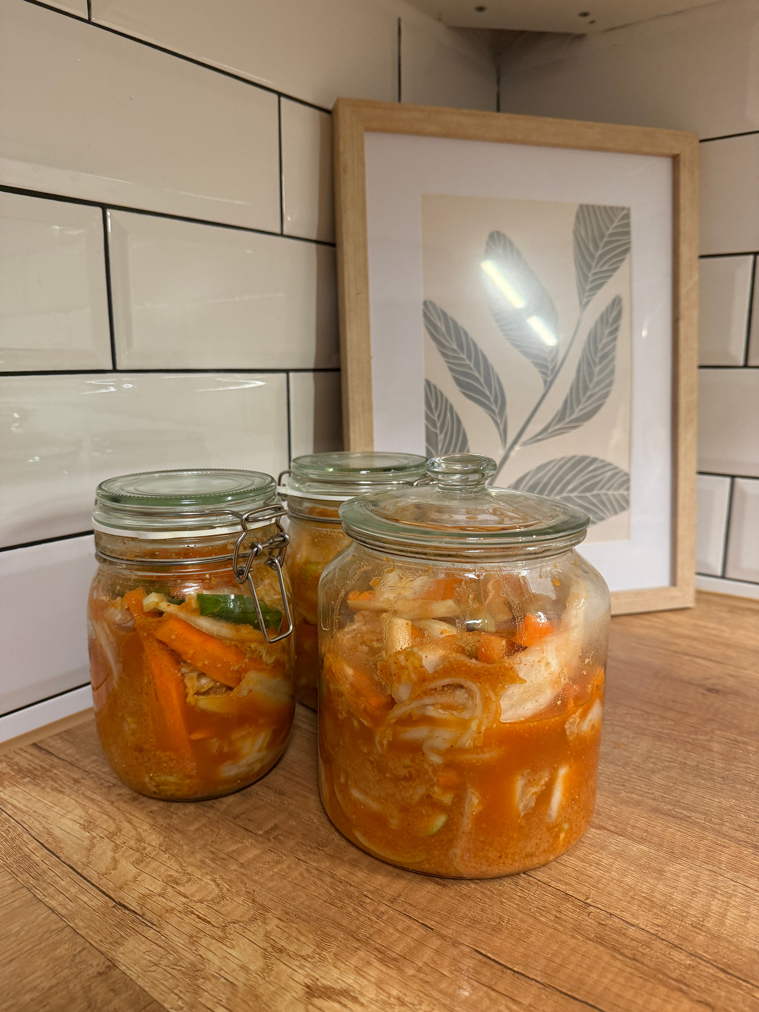 Drei Bügelgläser mit eingelegtem Kimchi aus Chinakohl, Karotten und Frühlingszwiebeln stehen auf einem Holztisch vor weißen Fliesen.