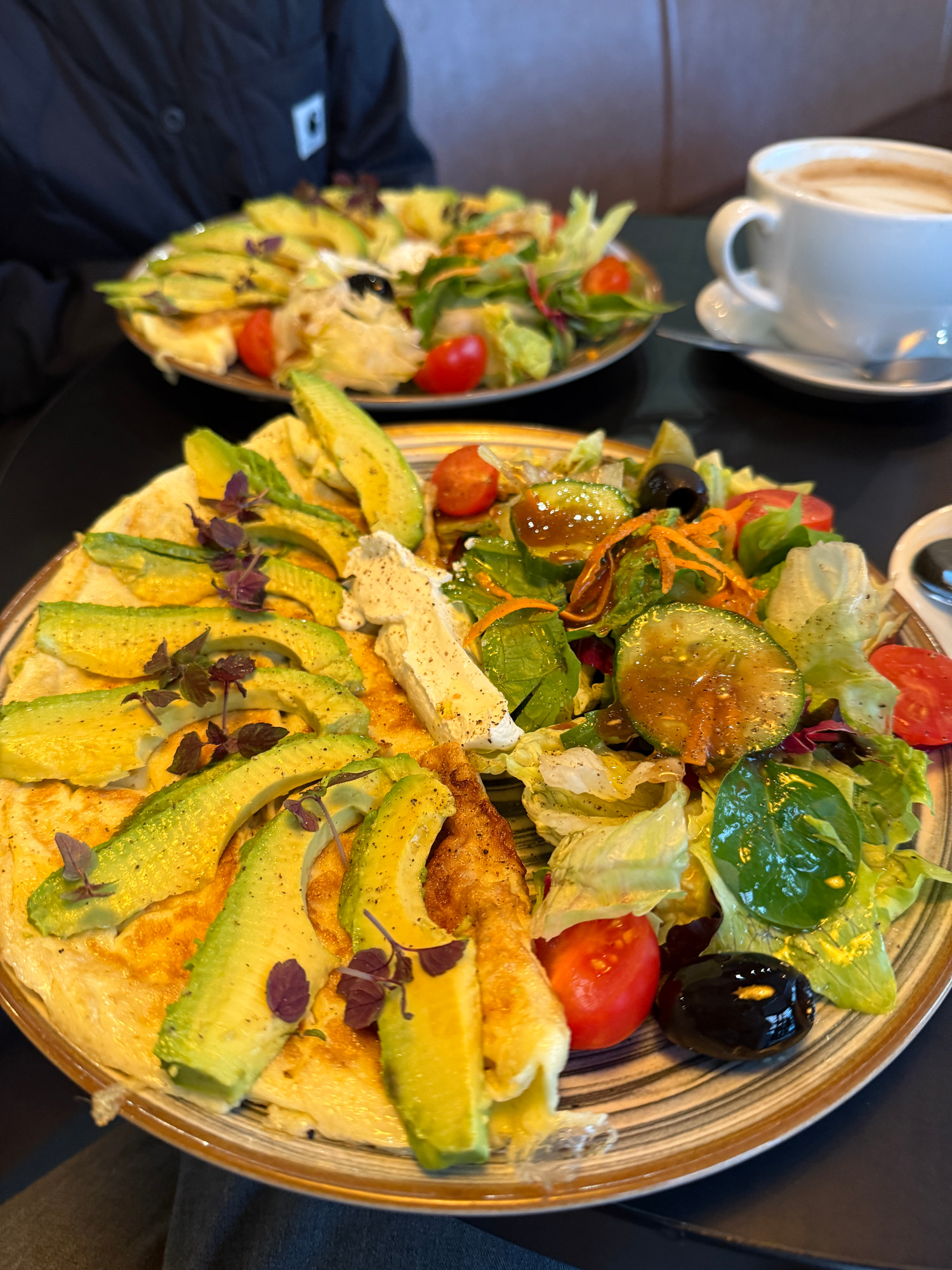 Ein Omelett mit fächerförmig aufgelegten Avocadoscheiben und Kräutern, daneben Blattsalat mit Tomaten, Oliven und Gurke auf einem dunklen Tisch.