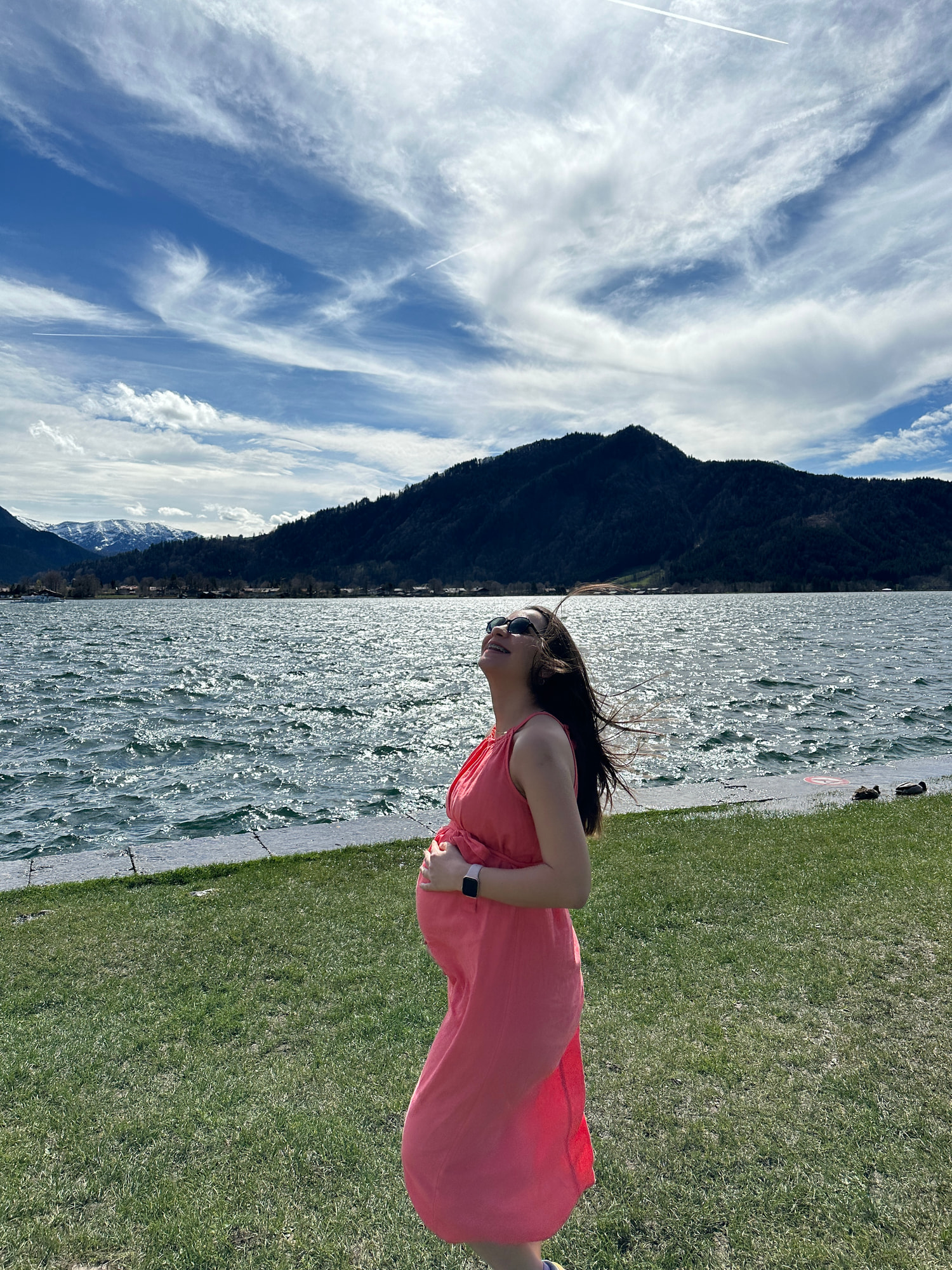 Elif Çimen Güngör steht schwanger in einem korallfarbenen Kleid auf einer Wiese am See und hält eine Hand an den Bauch, dahinter Berge und blauer Himmel.