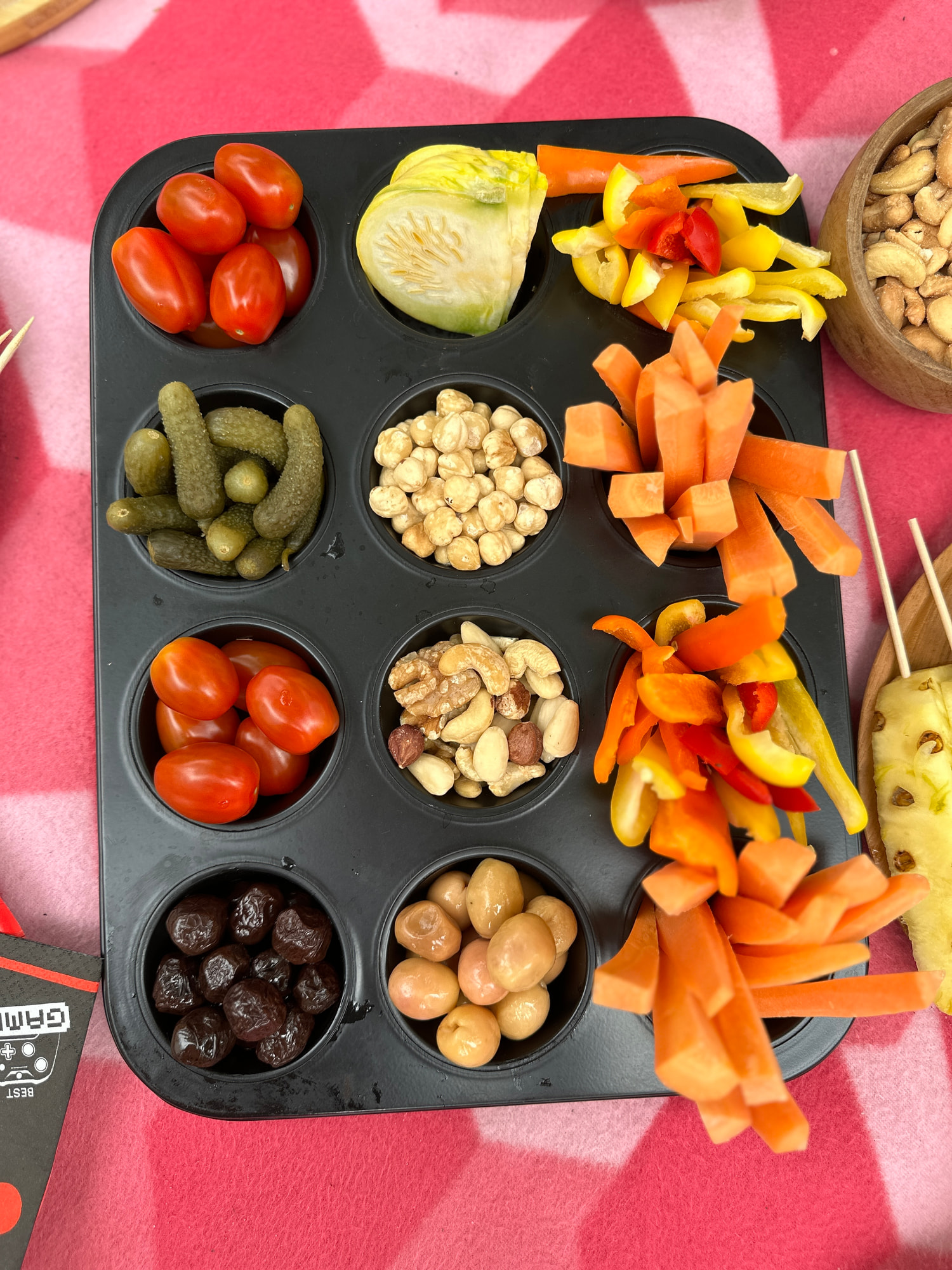 Eine Muffinform dient als Snackplatte mit Cocktailtomaten, Cornichons, Kichererbsen, Nüssen und Oliven, daneben Karotten- und Paprikastreifen auf einer rot gemusterten Decke.