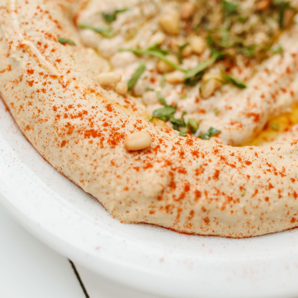 Nahaufnahme von cremigem Hummus auf weißem Teller, bestäubt mit Paprikapulver und garniert mit Pinienkernen, Petersilie und Olivenöl.